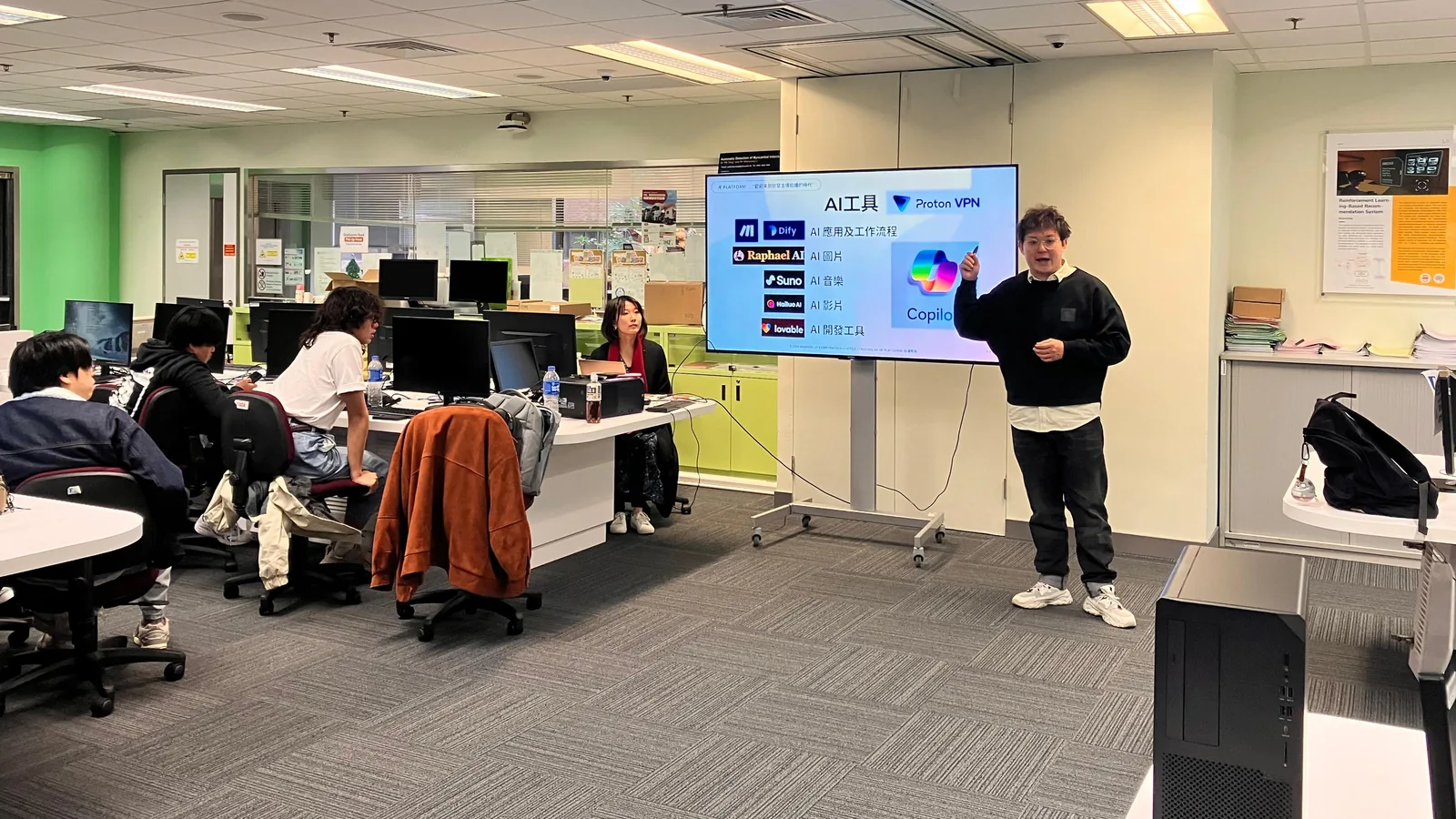 工作坊現場：導師站在投影螢幕旁講解，螢幕上列出當日介紹的 AI 工具，參加者在座位上各自使用自己的電腦